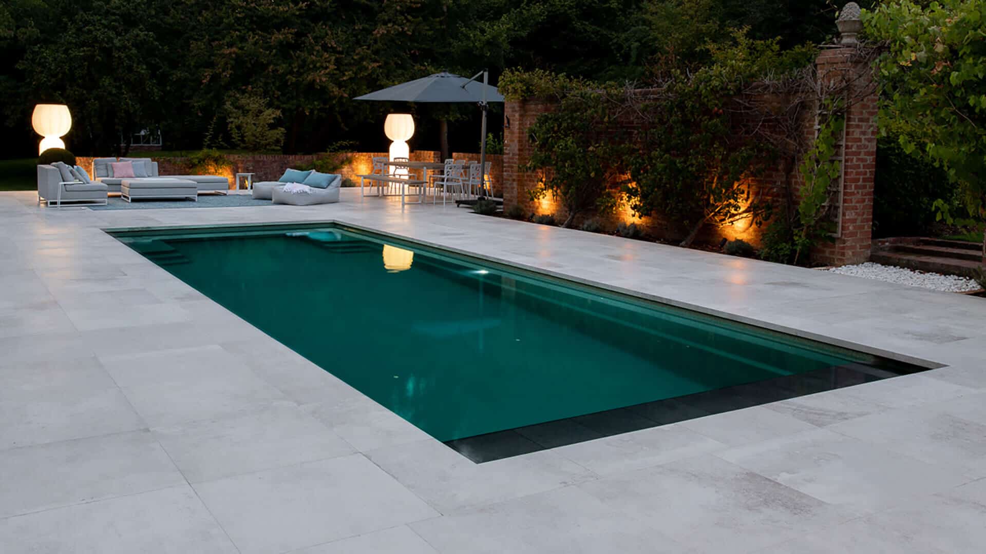 Evening view of a luxury outdoor swimming pool with soft garden lighting, modern patio furniture, and surrounding trees.
