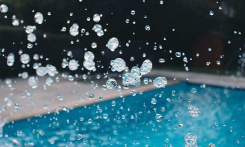 Close-up of water droplets splashing beside a bright blue outdoor swimming pool.