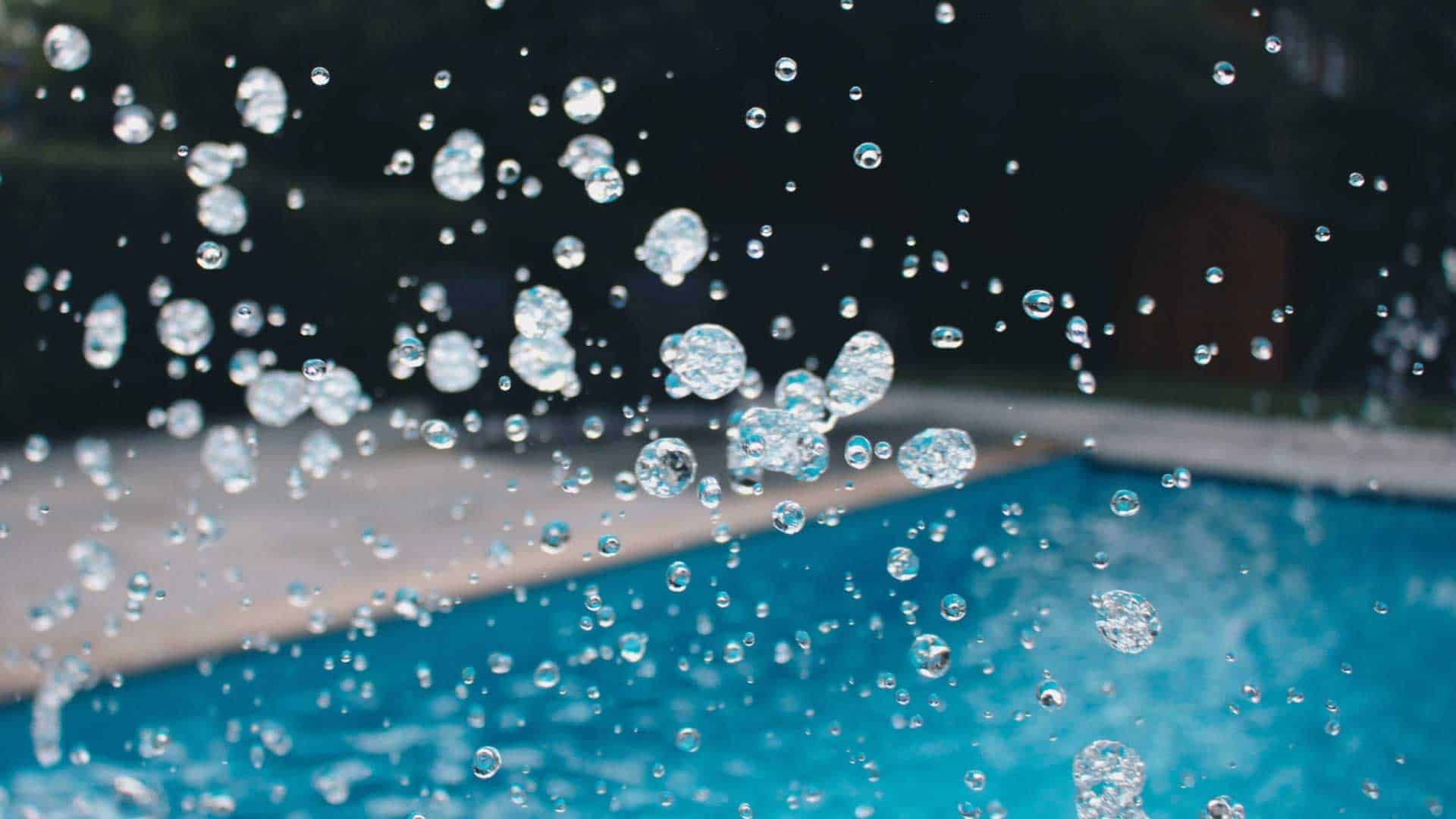 Close-up of water droplets splashing beside a bright blue outdoor swimming pool.