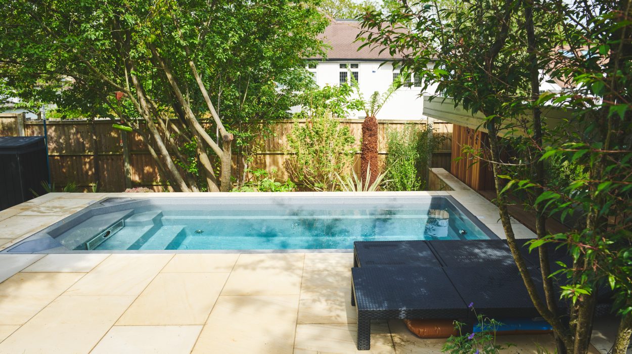 A modern back garden with a rectangular swim spa surrounded by trees and patio stones, featuring a lounge area with dark furniture.