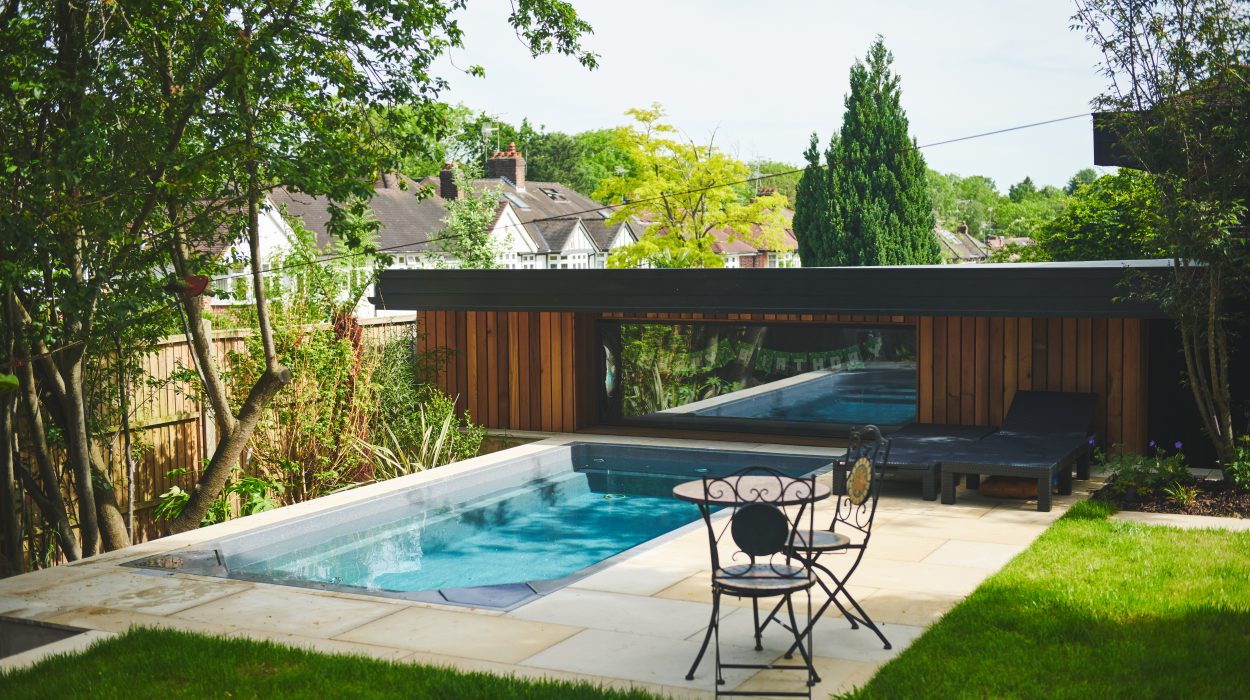 Modern garden with a compact swim spa surrounded by trees and a stylish timber-clad pool house with loungers.