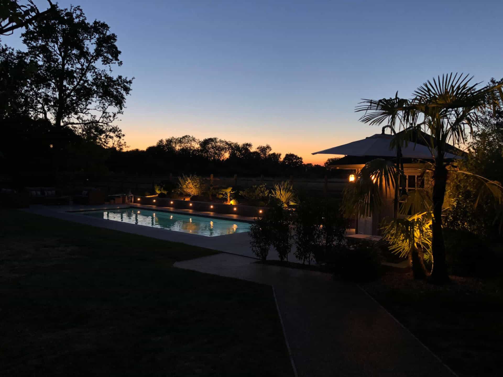 Outdoor swimming pool lit at sunset, with palm trees and garden lighting.