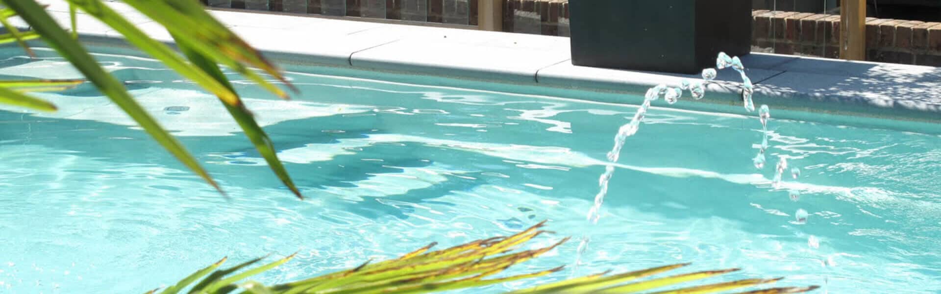 Modern garden pool with a gentle water feature surrounded by tropical plants and natural wood decking.