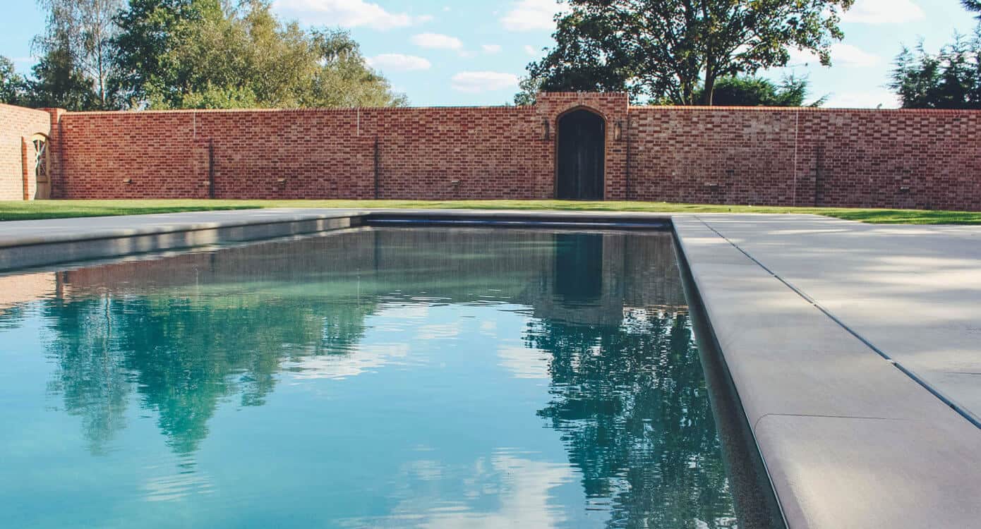 Outdoor swimming pool bordered by a red brick wall and surrounded by greenery in a private garden setting.