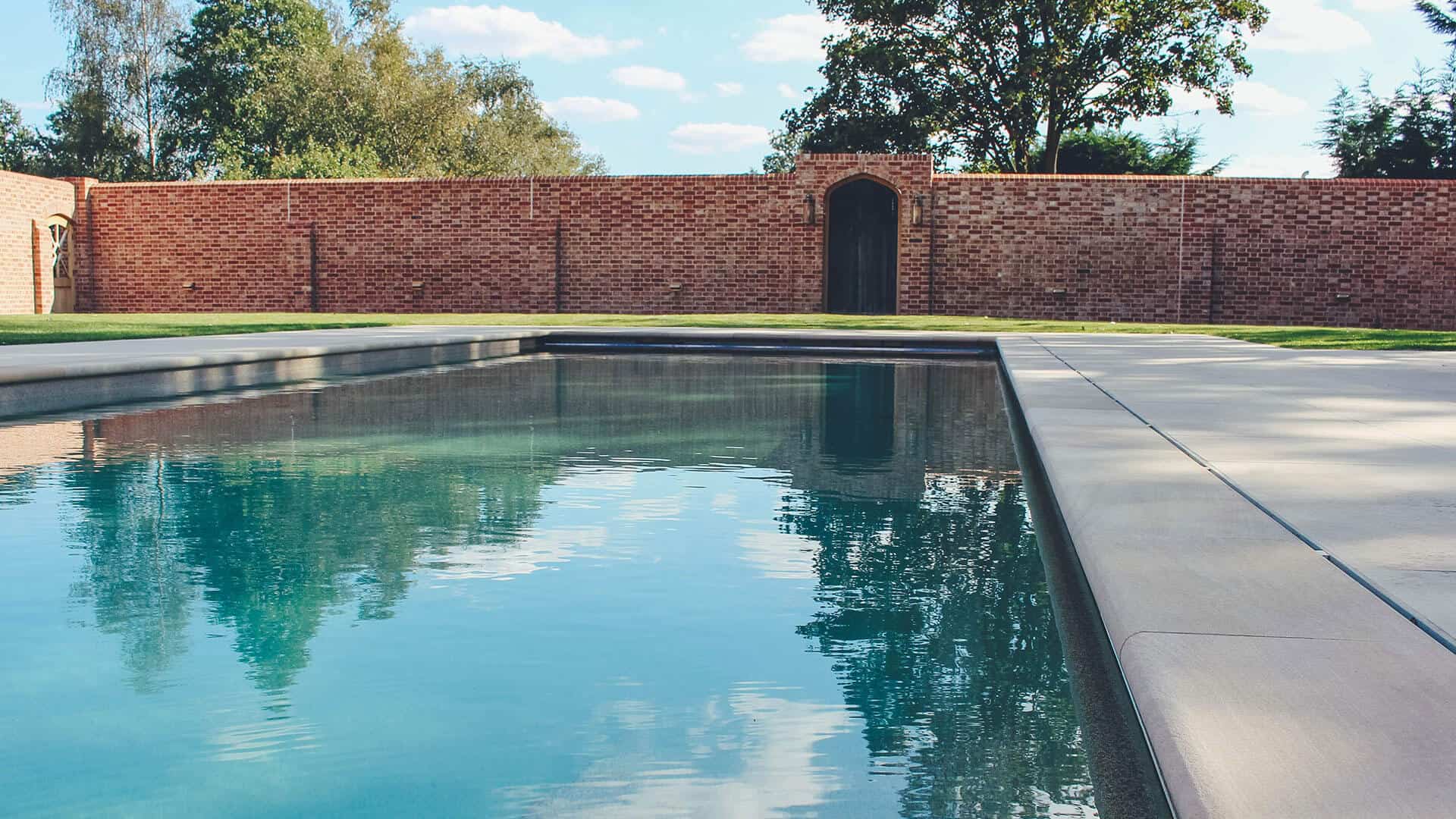 Outdoor swimming pool bordered by a red brick wall and surrounded by greenery in a private garden setting.